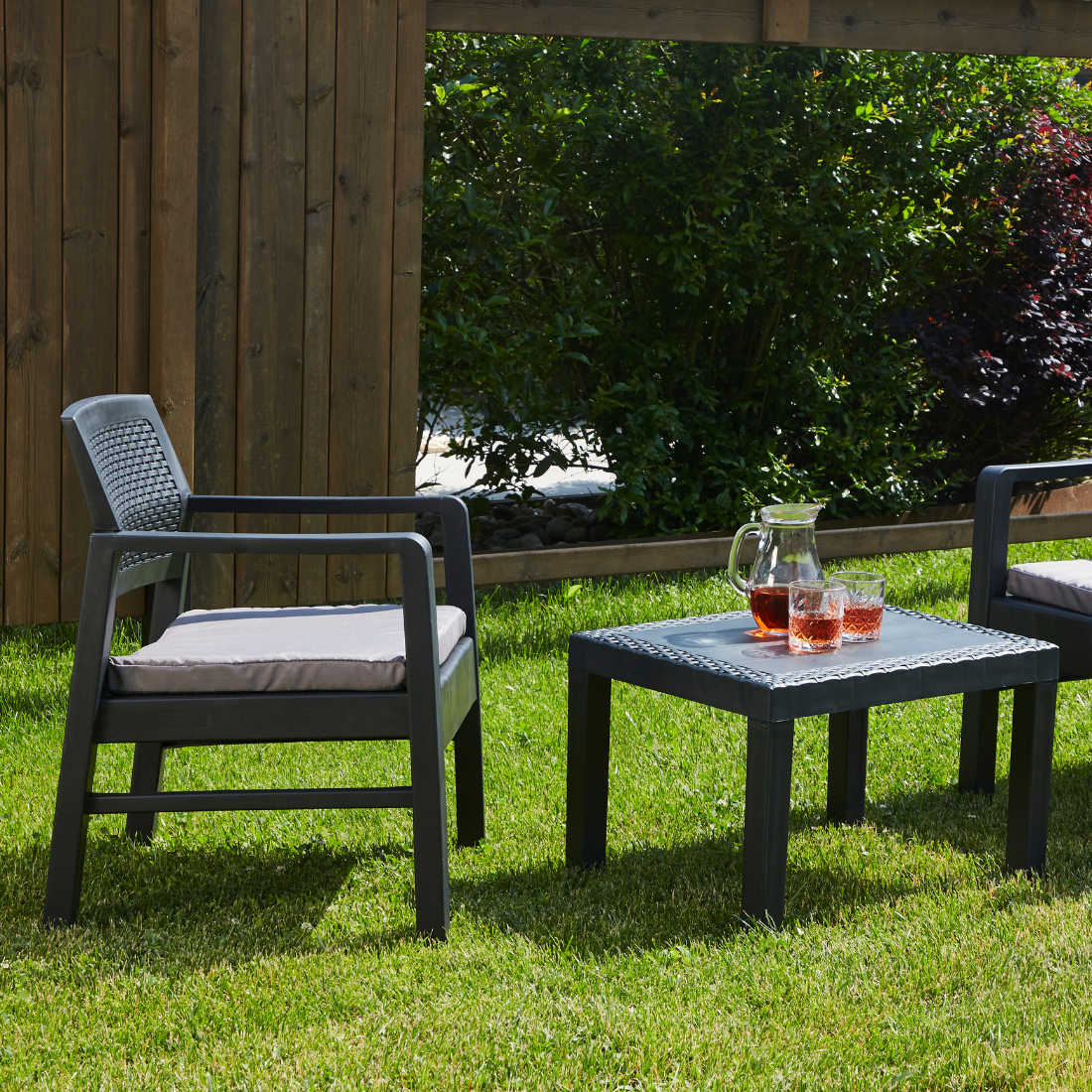 Conjunto de duas cadeiras baixas e uma mesa baixa quadrada IPAE PROGARDEN, da cor cinzenta, com almofada no assento e padrão entrançado. Exibido em ambiente exterior.