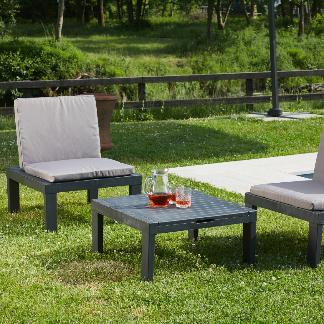 Conjunto IPAE PROGARDEN em plastico de cor cinzenta, composto por três módulos versáteis usado como duas cadeiras, com almofadas cinzentas no assento e no encosto e uma mesa. Exibido em ambiente exterior.