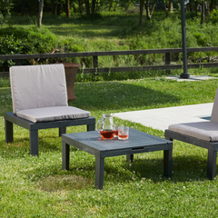 Conjunto IPAE PROGARDEN em plastico de cor cinzenta, composto por três módulos versáteis usado como duas cadeiras, com almofadas cinzentas no assento e no encosto e uma mesa. Exibido em ambiente exterior.