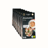 Ensaladilla em embalagem de cartão e plástico transparente, com pedaços de batata, cenoura, ervilhas e maionese. A embalagem tem um rótulo verde com o nome do produto e informações nutricionais. Fundo branco.