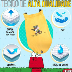 Informação sobre o tecido de um colete  Zooz para cão na cor amarela, com detalhes em preto, com estampa do  personagem a deslizar na neve com um gorro e cachecol, acompanhado do Woodstock. Vista frontal, sobre fundo branco. 