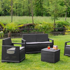 Conjunto IPAE PROGARDEN, composto por um sofá de dois lugares, dois cadeirões e uma mesa, em plástico com acabamento trançado e almoçadas de acento de cor cinzento. Exibido em ambiente exterior. 