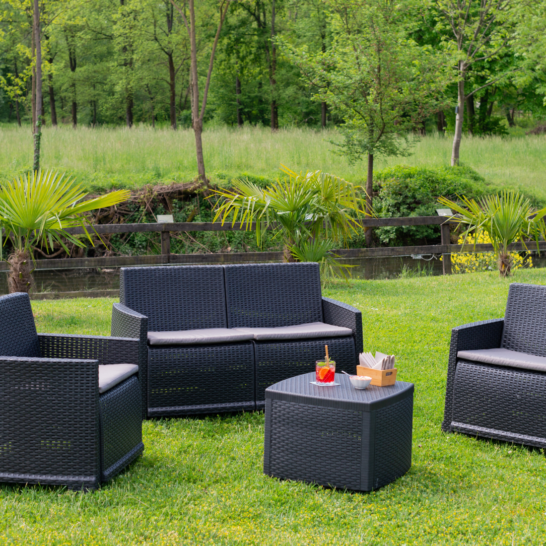 Conjunto de dois cadeirões, um cadeirão duplo e uma mesa baixa, com acabamento entrançado, de cor cinzenta, almofada no assento e  com braços de apoio. Exibido em ambiente exterior. 