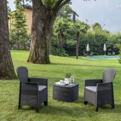 Conjunto de 2 cadeirões e uma mesa baixa IPAE PROGARDEN, de cor cinzenta, com acabamento entrançado e assento com almofada da cor bege. Exibido em ambiente exterior. 